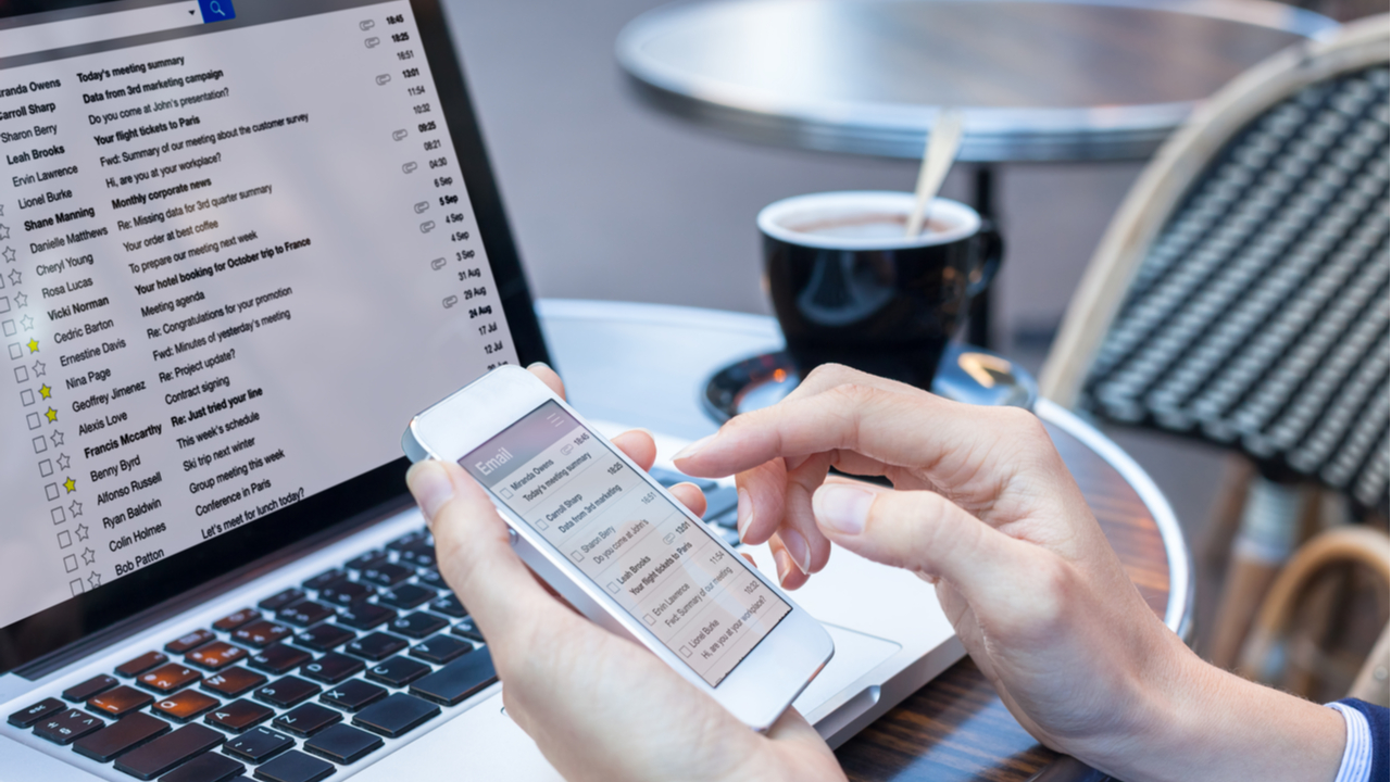 Person at desk checking email on smartphone and laptop.